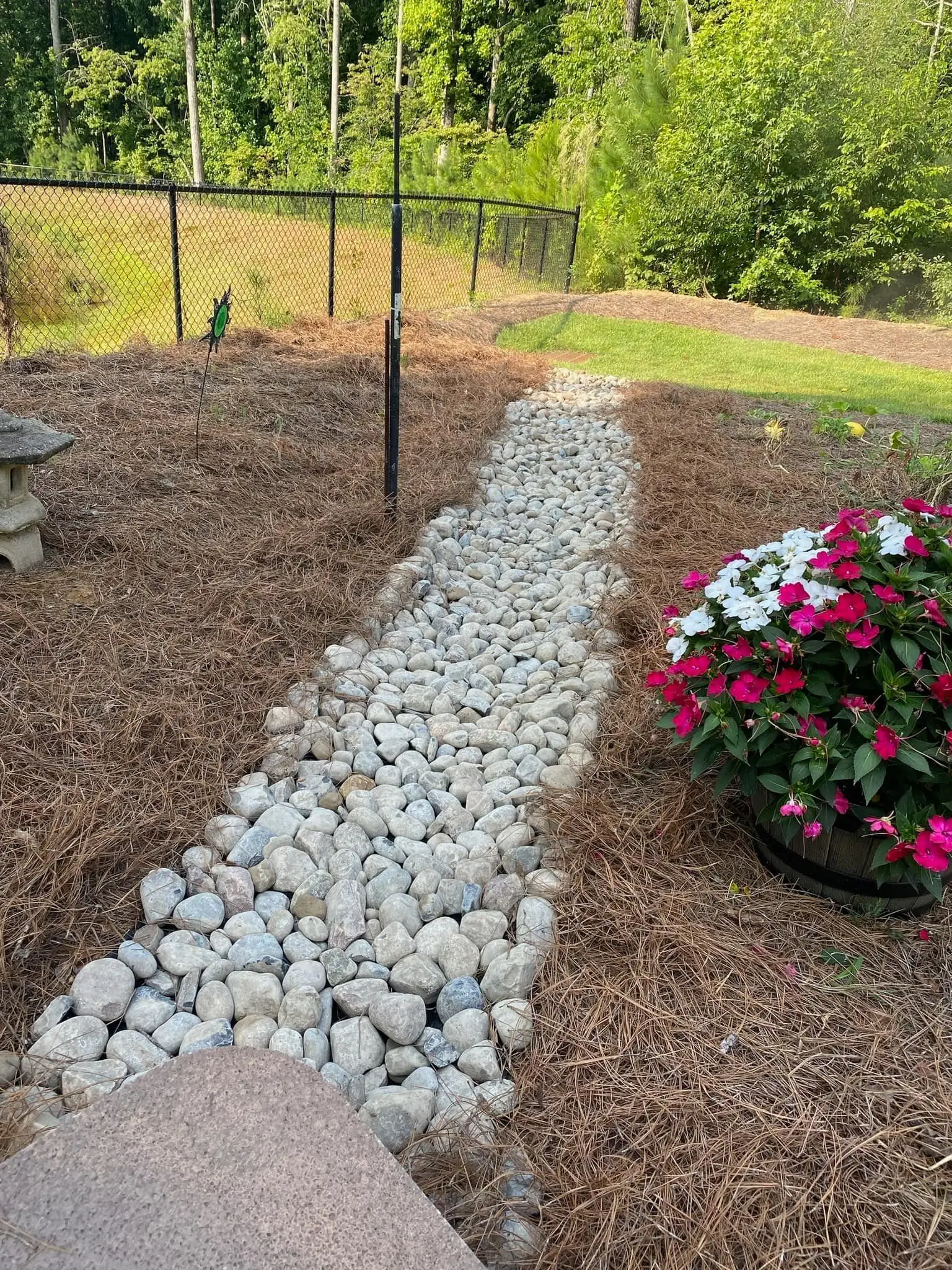 River-rock drainage channel running through a pine-straw bed beside flowering planters