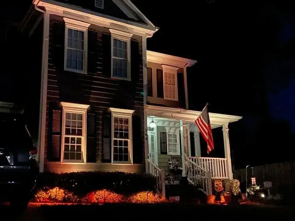 Two-story house with porch, American flag, and orange lights illuminating the yard and facade at night.